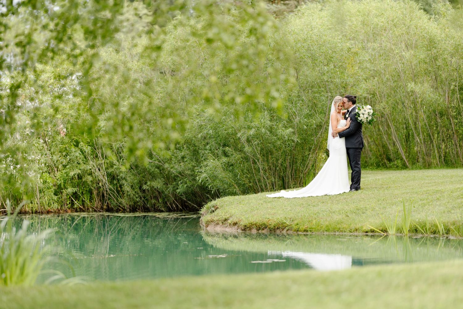 A couple in wedding attire embraces on a lush green golf course beside a tranquil pond.