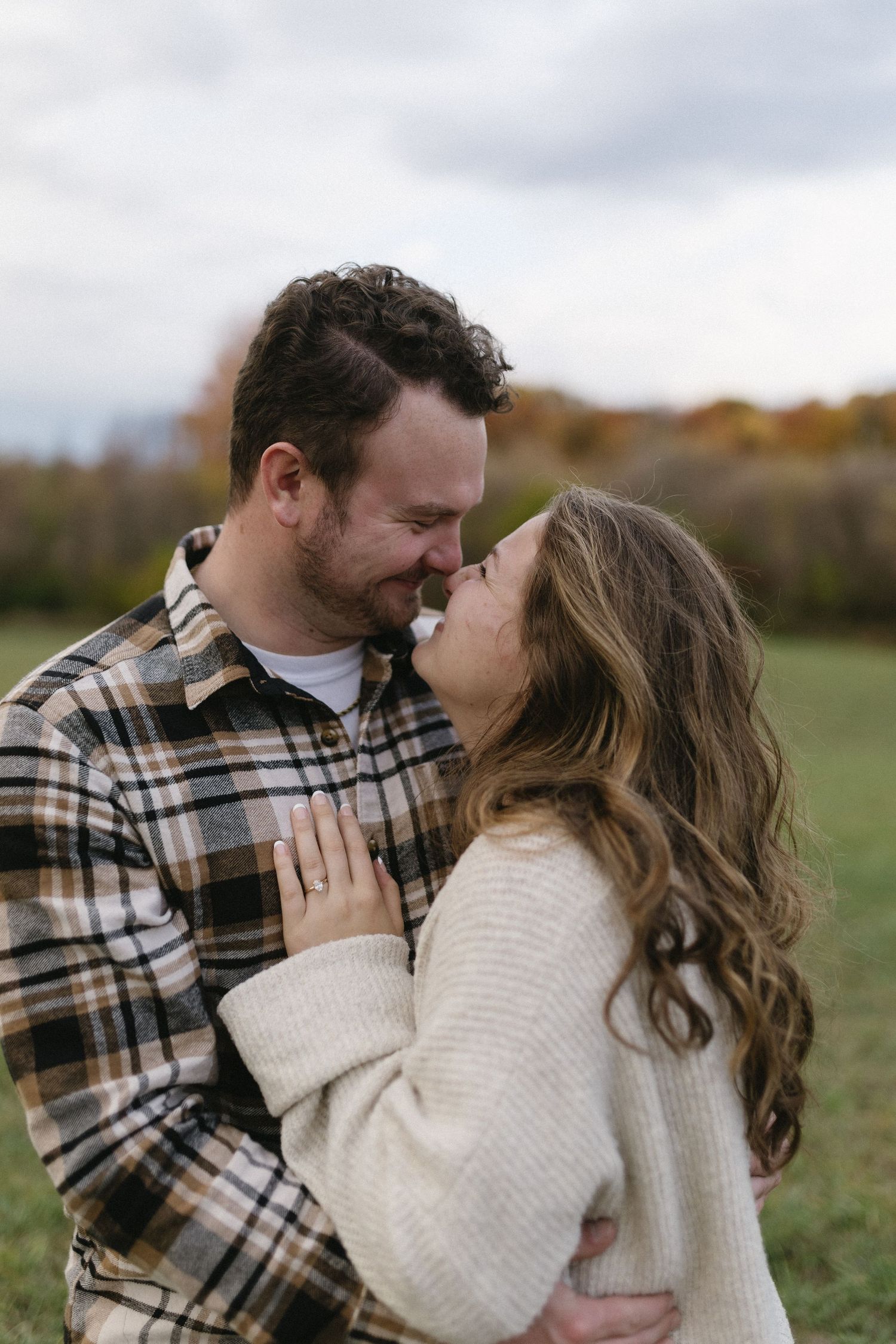 A romantic couple embraces outdoors in autumn wearing cozy sweaters and plaid against a nature backdrop.