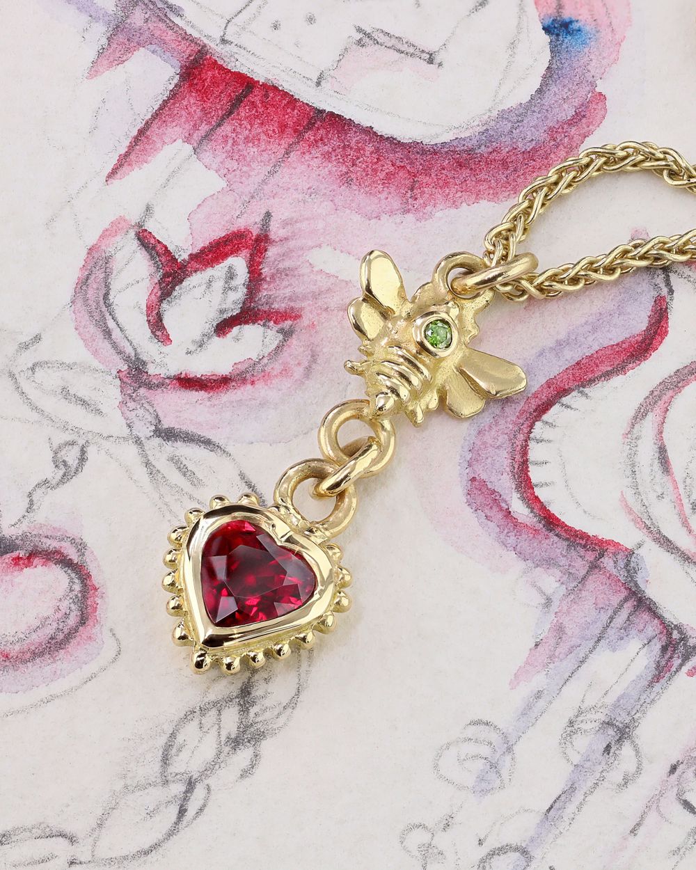 Gold necklace pendant featuring red heart crystal and decorative bee charm on floral background.