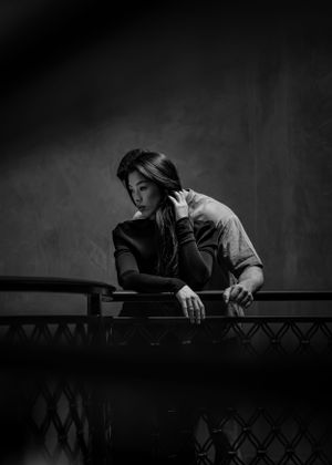A moody black and white portrait shows someone leaning thoughtfully against a decorative railing in dramatic lighting.