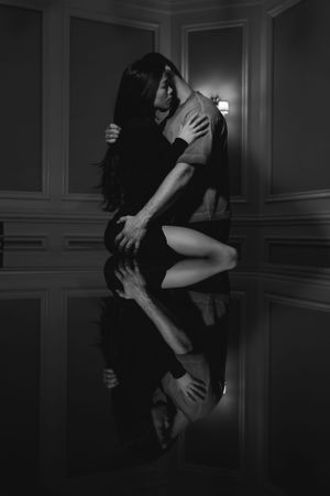 Artistic black and white couple's boudoir portrait of a couple embracing in a dimly lit room with dramatic mirror reflection on floor.