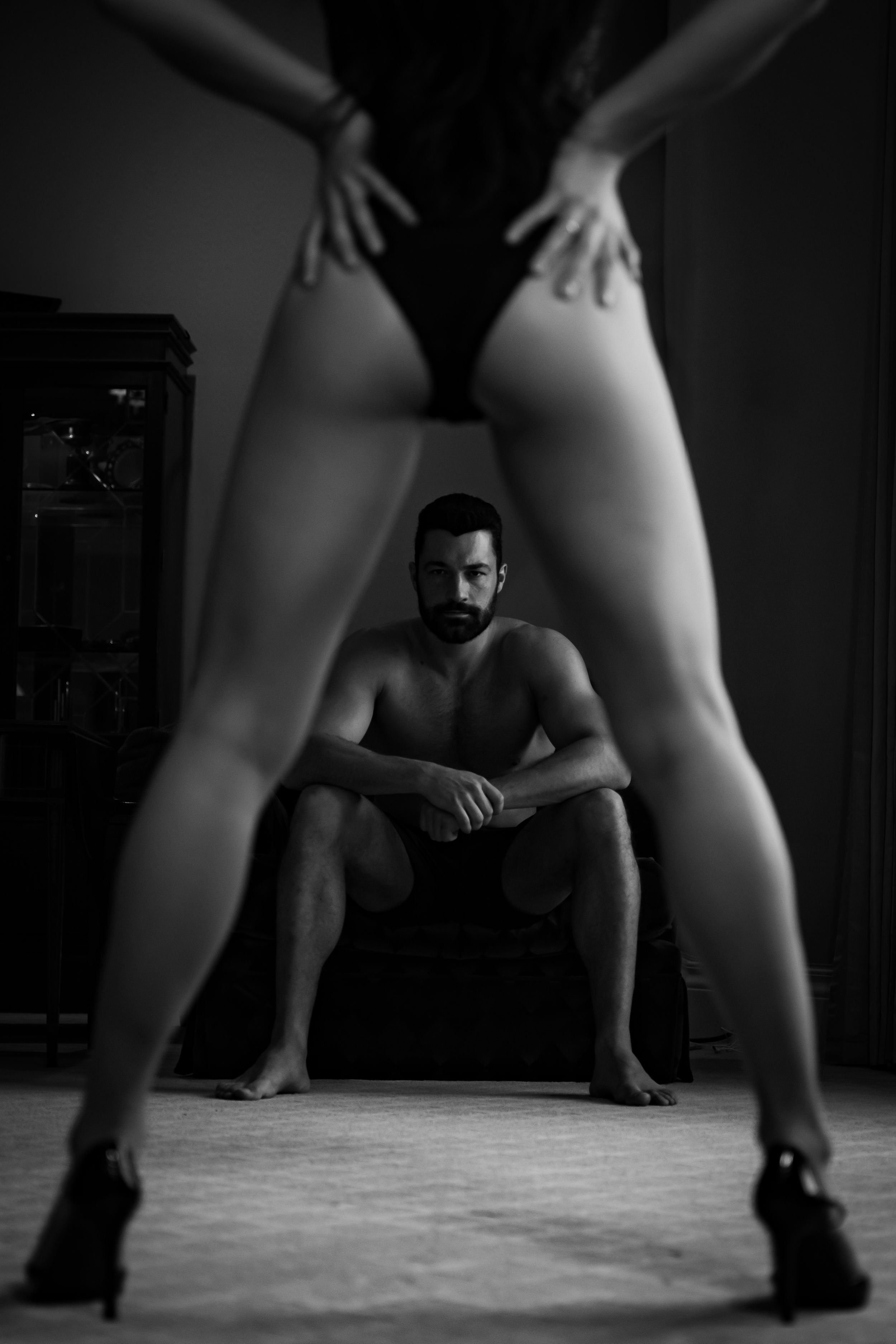Artistic black and white couple's boudoir photo with female silhouette in heels and figure seated on floor in dark setting.