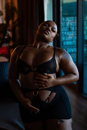 Moody boudoir photography with dramatic window lighting on black model in black lingerie.