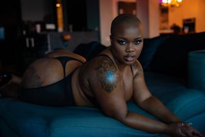A black woman in moody lighting lies on a blue velvet couch wearing black lingerie with a shoulder tattoo visible.