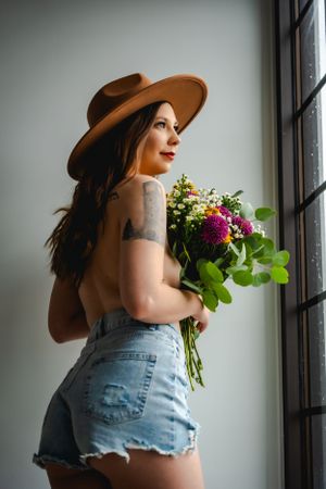 A woman in a hat and denim shorts holds a bouquet of flowers covering her bare chest while near a window at The White Space Las Vegas.