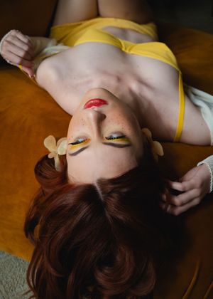 Artistic beauty portrait of someone wearing yellow top with vibrant eye makeup and red lipstick lying on orange surface.
