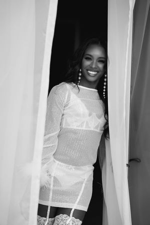 Black and white portrait of person wearing sheer white corset dress and lace stockings while posing in doorway.