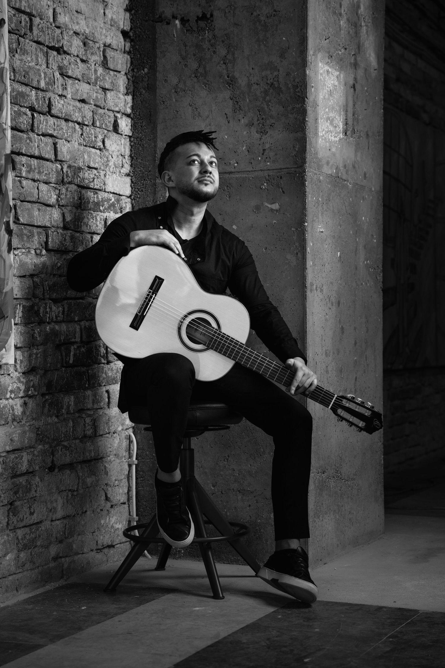 Black and white portrait of musician with acoustic guitar sitting against brick wall in urban setting.