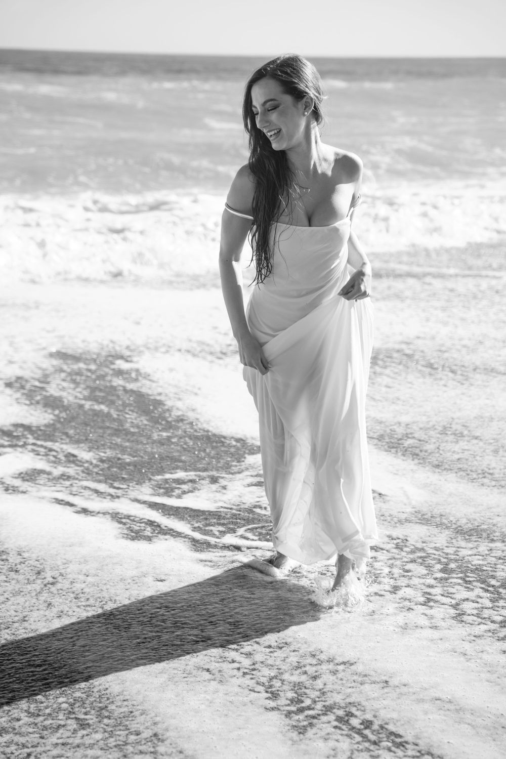 Elegant woman in flowing white dress walks along ocean shore in black and white.