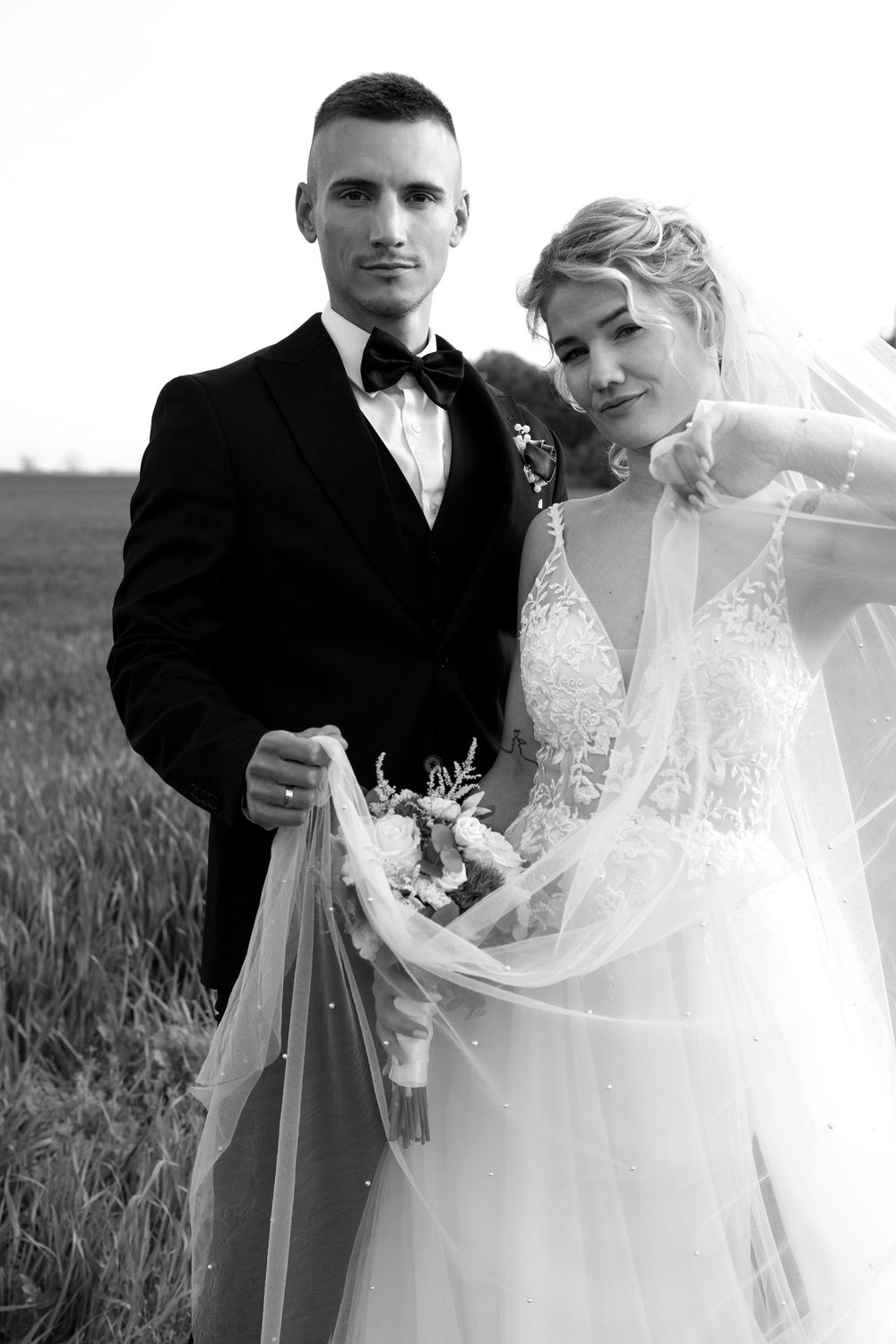 Elegant black and white wedding portrait with couple holding veil in field.