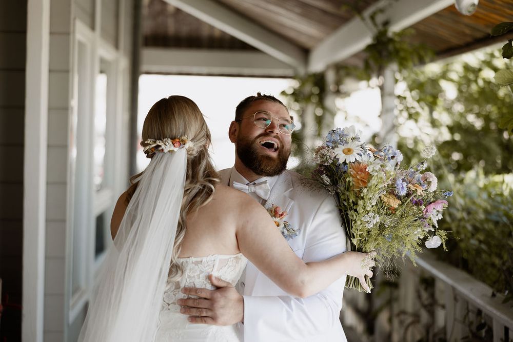 A joyful moment between newlyweds on a porch with the bride holding a wildflower bouquet.