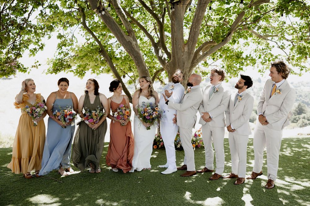 Wedding party members seated in row on grassy lawn under tree shade wearing coordinated dresses and attire.