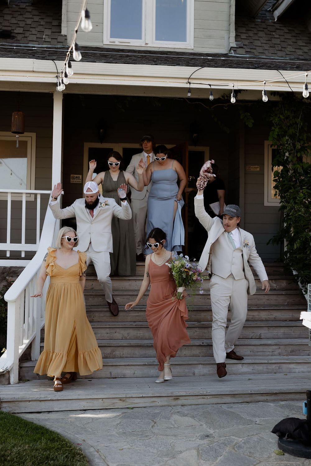Wedding party members joyfully dancing and jumping off steps in front of a gray house wearing vintage-inspired outfits.