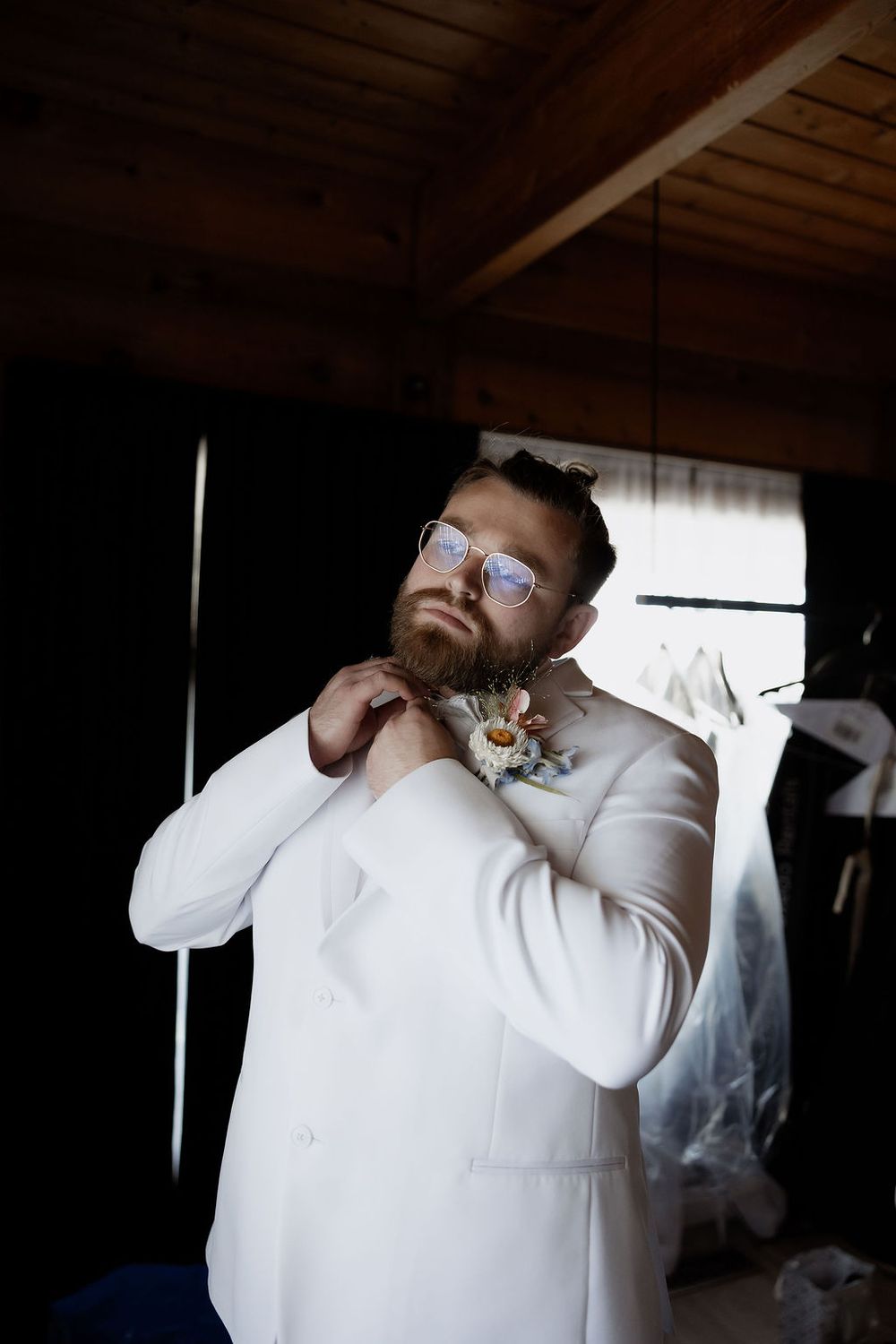 A person in a white suit jacket and glasses adjusts their tie while getting ready in a dimly lit room.