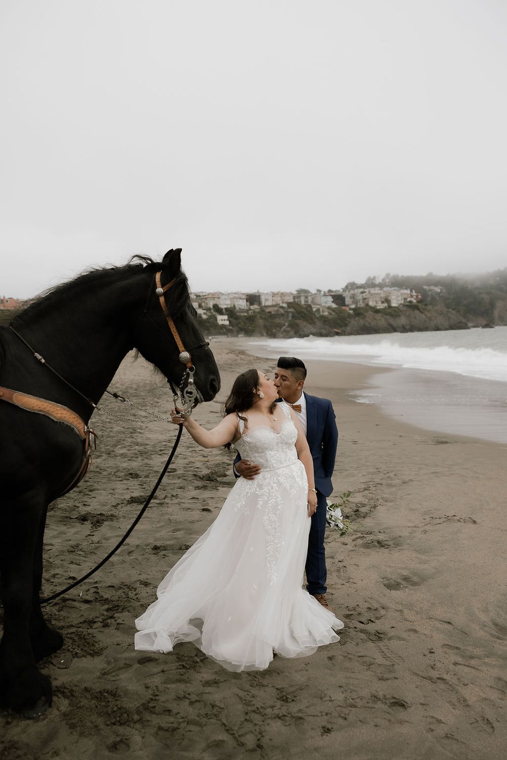 A romantic beach wedding photoshoot with a couple and their black horse on a misty coastline.