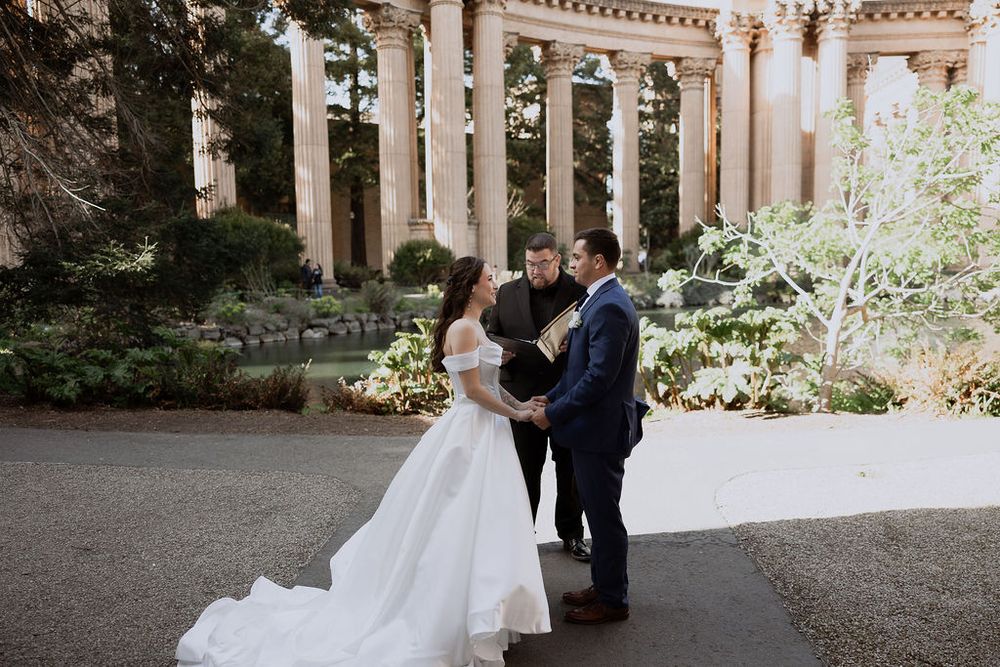 A couple in wedding attire stands on a scenic path surrounded by lush greenery and historic architecture.