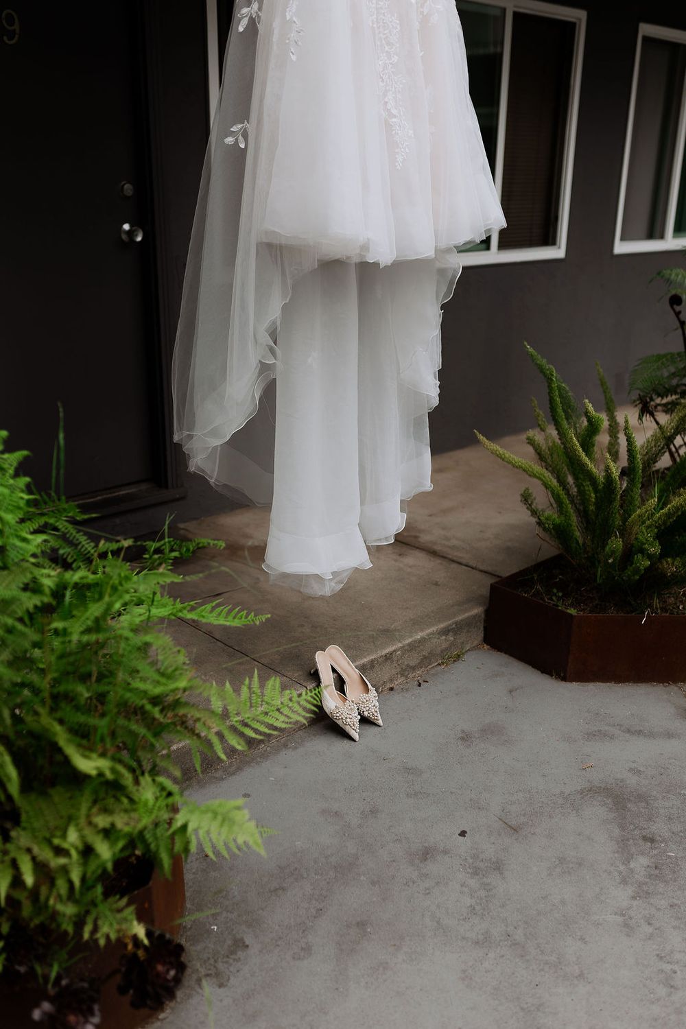 A white wedding dress and heels are photographed with green ferns against a gray concrete porch.
