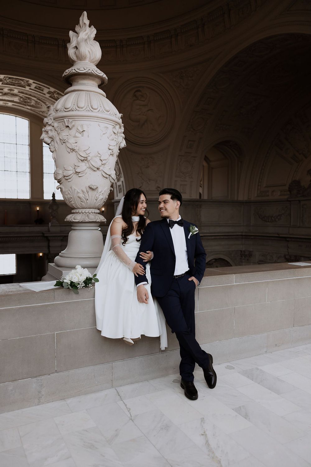 An elegant wedding couple sits together on ornate white marble steps in a grand architectural setting with decorative details.