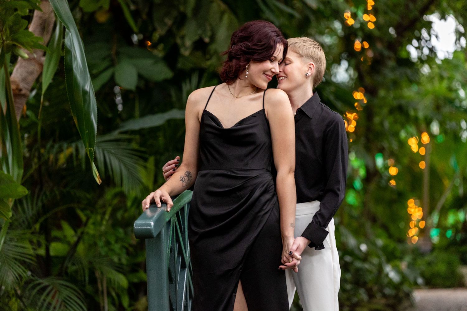 Two women in elegant attire share a tender moment surrounded by tropical greenery.