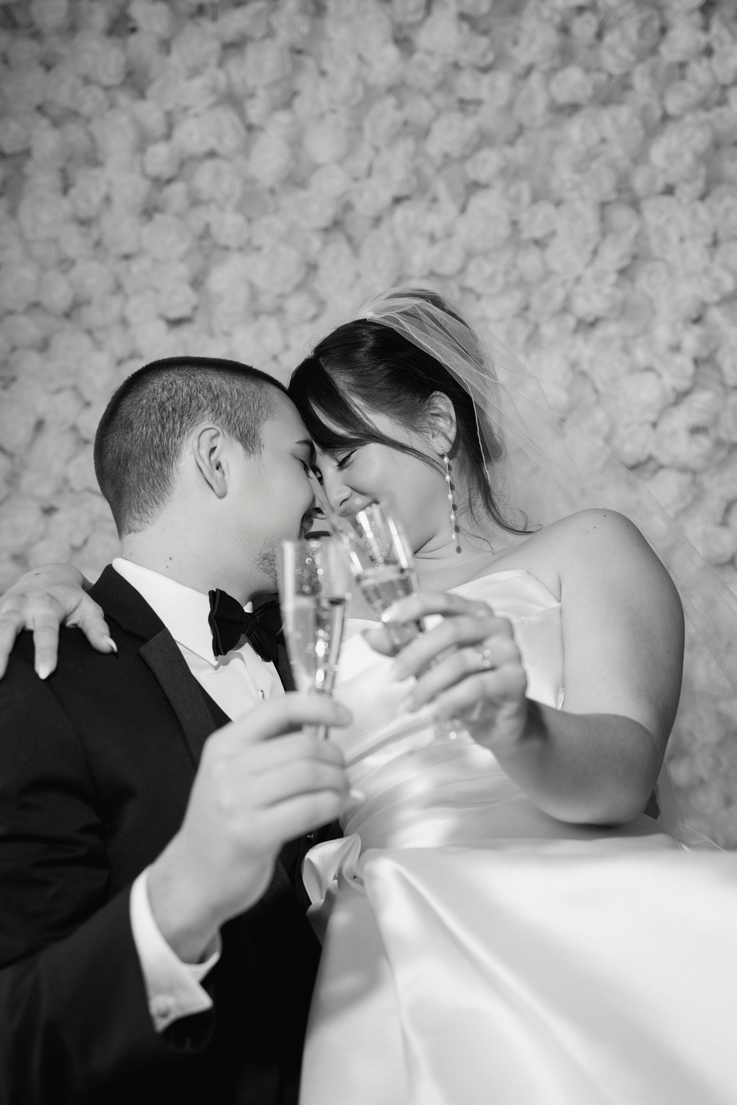 Black and white photo of a bride and groom toasting with champagne glasses, smiling closely together before a floral wall.