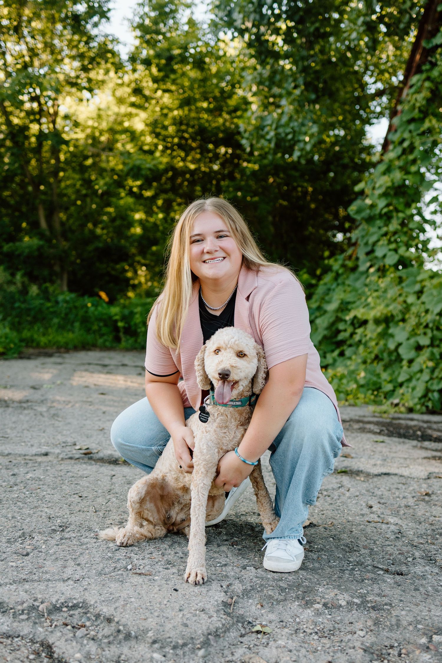 Smiling blonde woman crouching outdoors with a fluffy golden doodle dog on a paved surface.