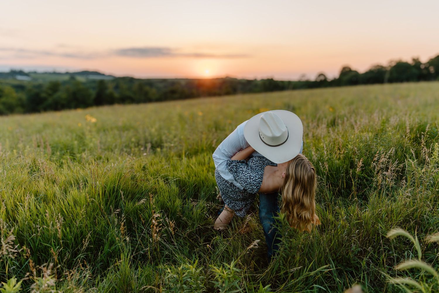 Couple shares a tender embrace in a lush green field during a golden sunset.