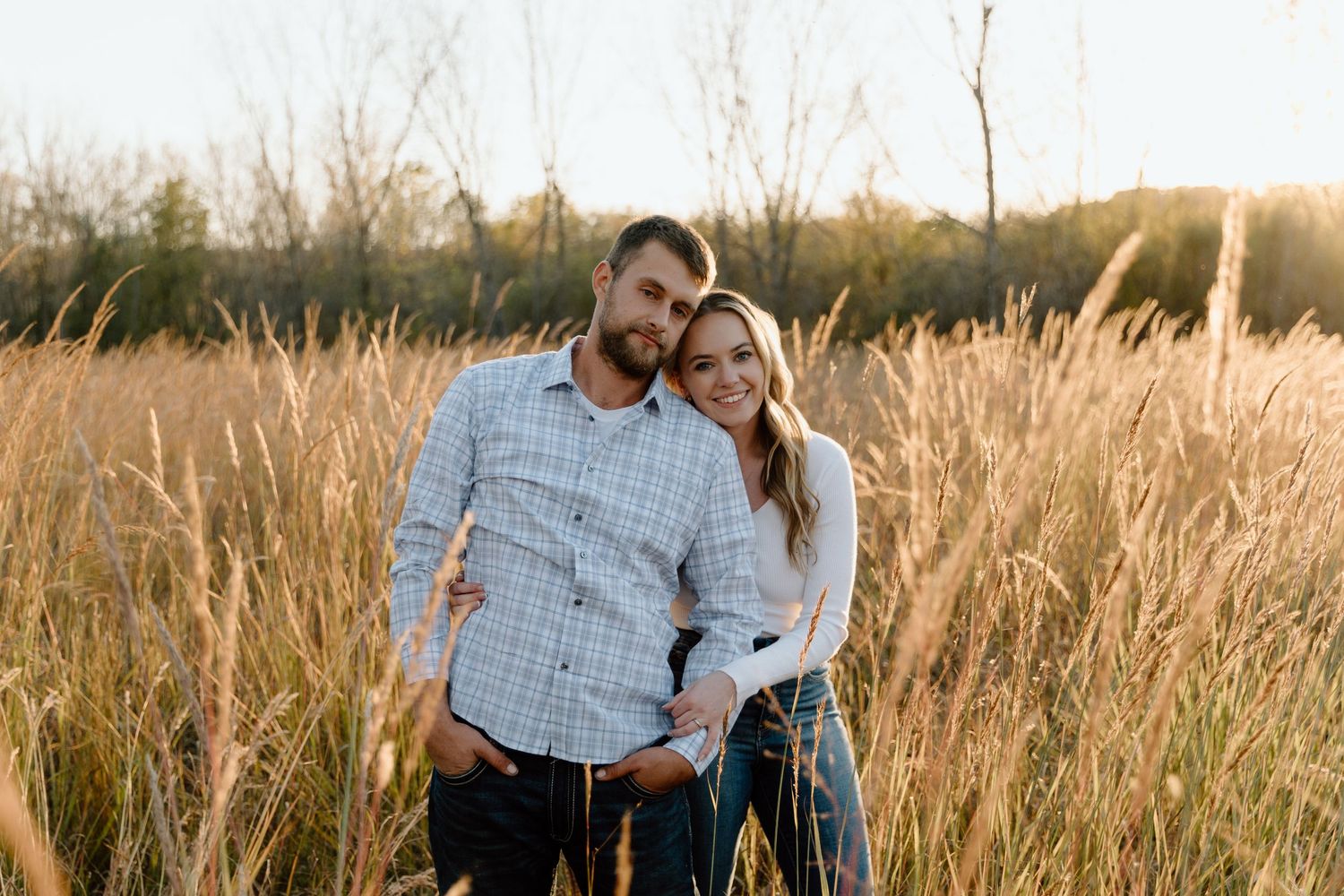 Couple smiling together in golden tall grass field at sunset, man in plaid shirt embracing woman in white top.