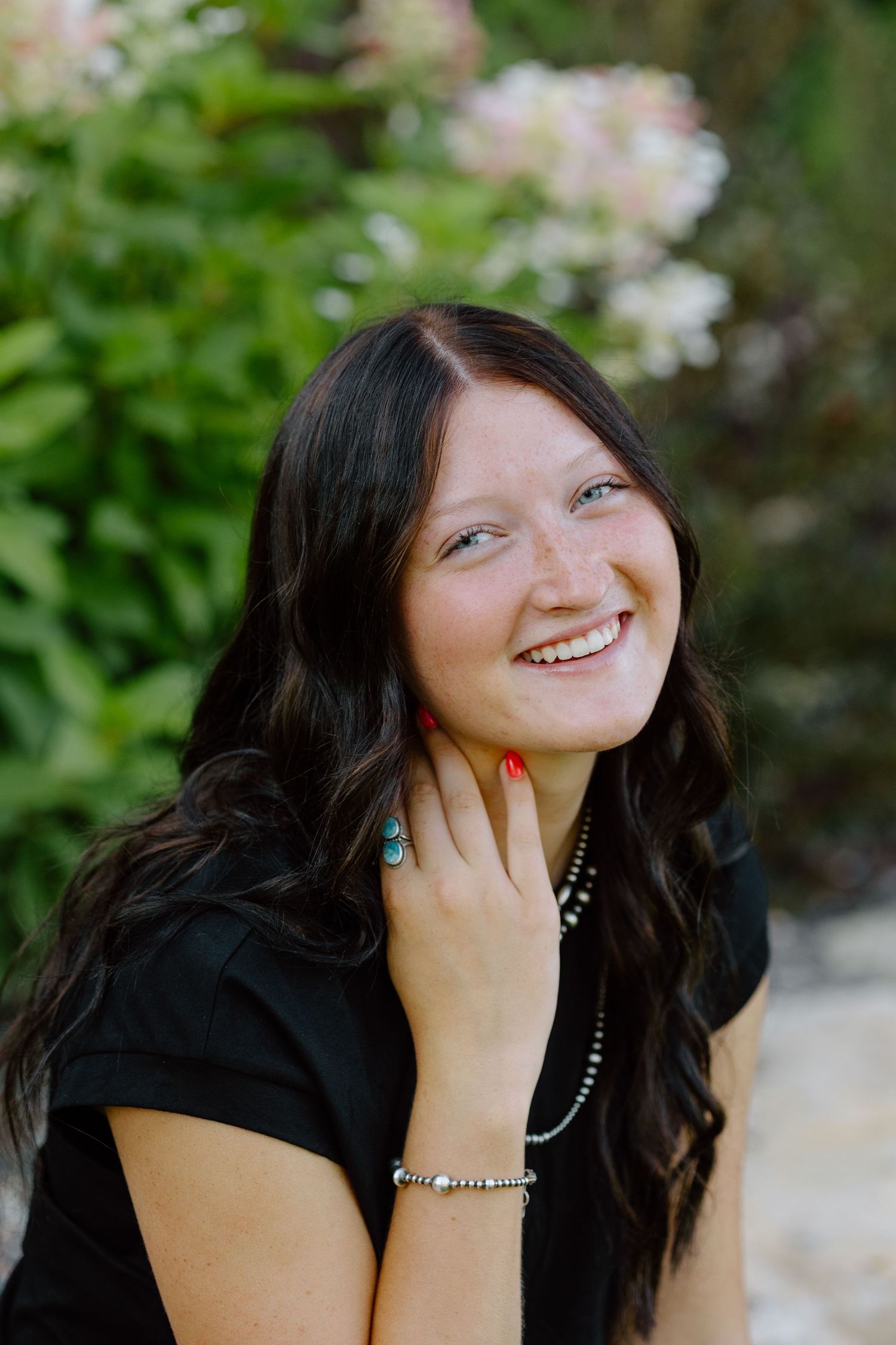 Young woman with dark wavy hair smiling outdoors near blooming flowers, wearing black top and turquoise jewelry.