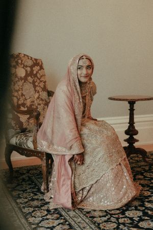 A traditional bride in a pink and gold embroidered dress with matching hijab sits elegantly on an antique chair.