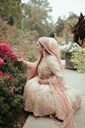 A bride in a cream embroidered dress and pink hijab sits by blooming garden flowers.