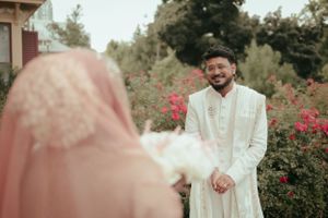 Muslim couple in traditional wedding attire stand among blooming rose bushes in a garden setting.
