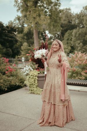 Woman in ornate peach-colored traditional dress holds white bouquet in garden setting.