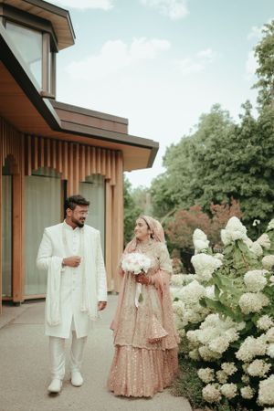 Couple in traditional wedding attire walk together past blooming white hydrangea bushes.