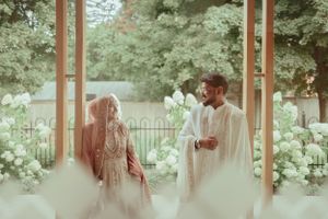 Couple in traditional dress share intimate moment in garden setting framed by white flowers.