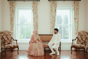 Couple in traditional attire seated in elegant room with floral curtains and antique chairs.
