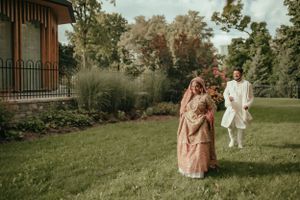 Couple in traditional South Asian wedding attire posing in a garden with historic building backdrop.