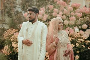 Elegant couple in traditional wedding attire standing among blooming pink roses in a garden.