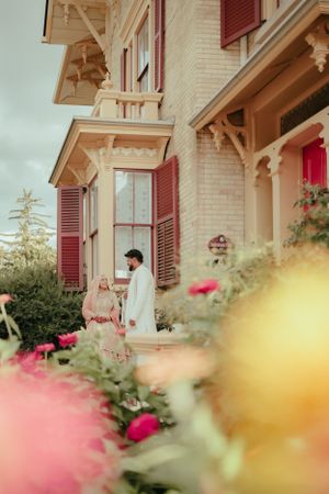 Traditional wedding couple posed at Victorian-style house with red shutters and blooming flowers.