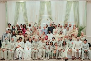 Large wedding gathering wearing coordinated cream and white outfits seated together in an elegant hall with green curtains.