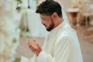 Person in white embroidered formal attire praying during ceremony.