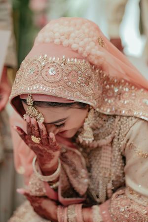 Bride in peach embroidered attire and traditional jewelry covering face with hennaed hands.