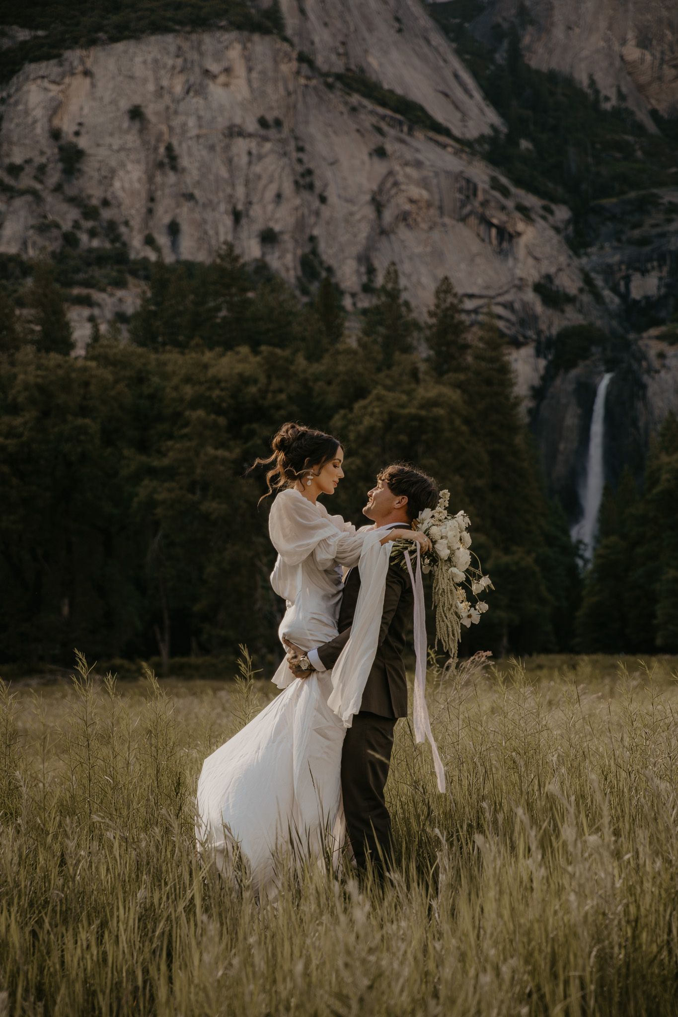 A romantic wedding photo of a couple embracing in a meadow beneath towering cliffs and a waterfall.
