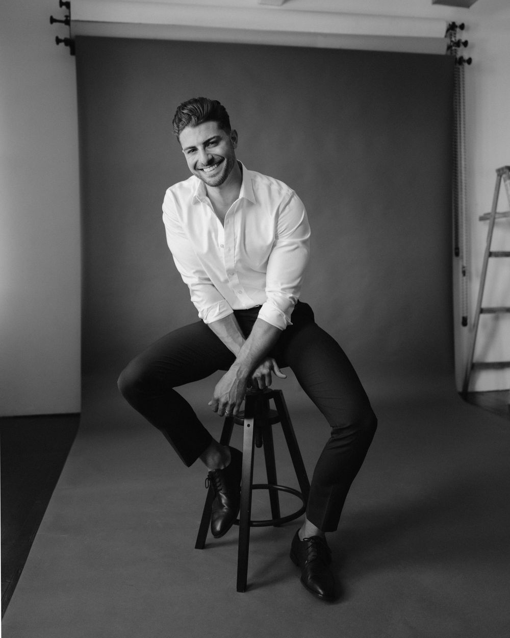 Black and white studio portrait of a person in a white shirt and dark pants sitting casually on a wooden stool.