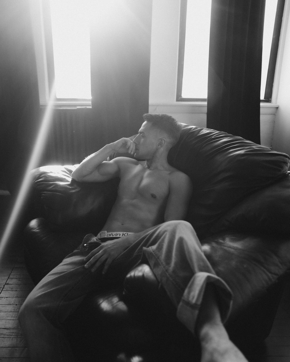 Moody black and white photo of shirtless person reclining on dark couch in sunlight.