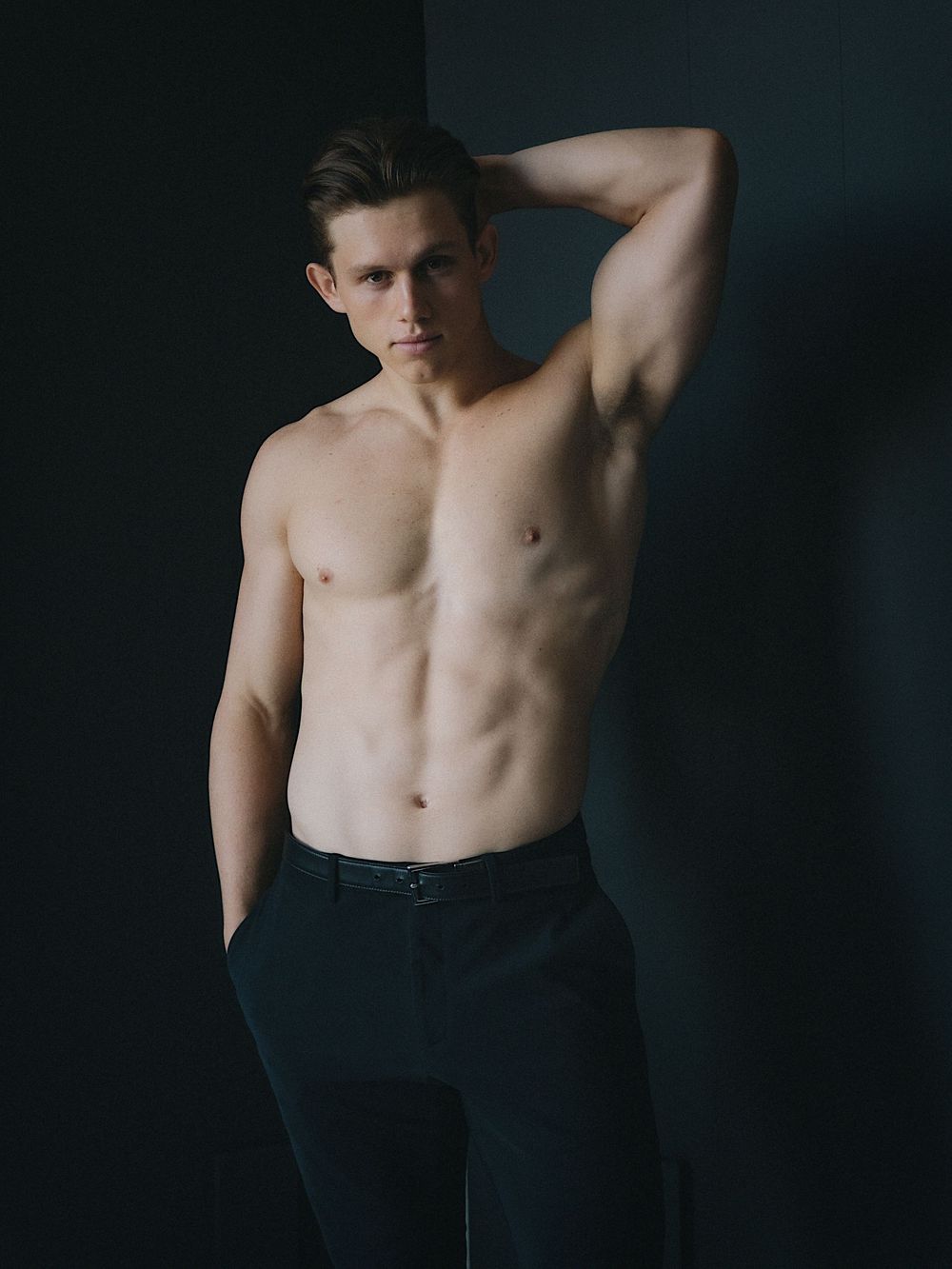 Fitness model poses shirtless against dark background showcasing athletic physique.