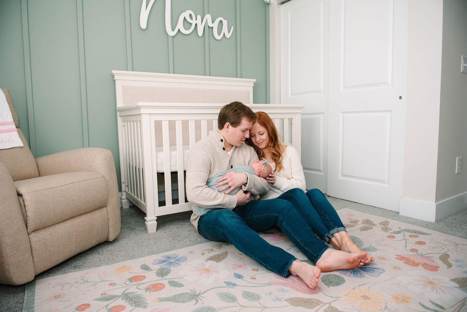 A dog rests on a bed next to someone feeding a baby in a soft, natural light setting.
