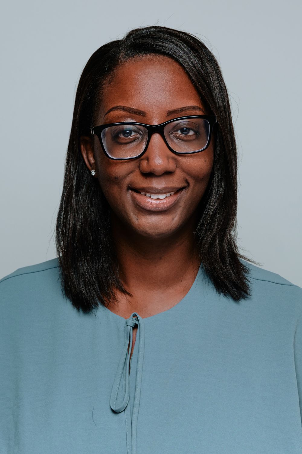 Person wearing glasses and a gray zip-up sweater smiles warmly against a light blue background in a headshot series.