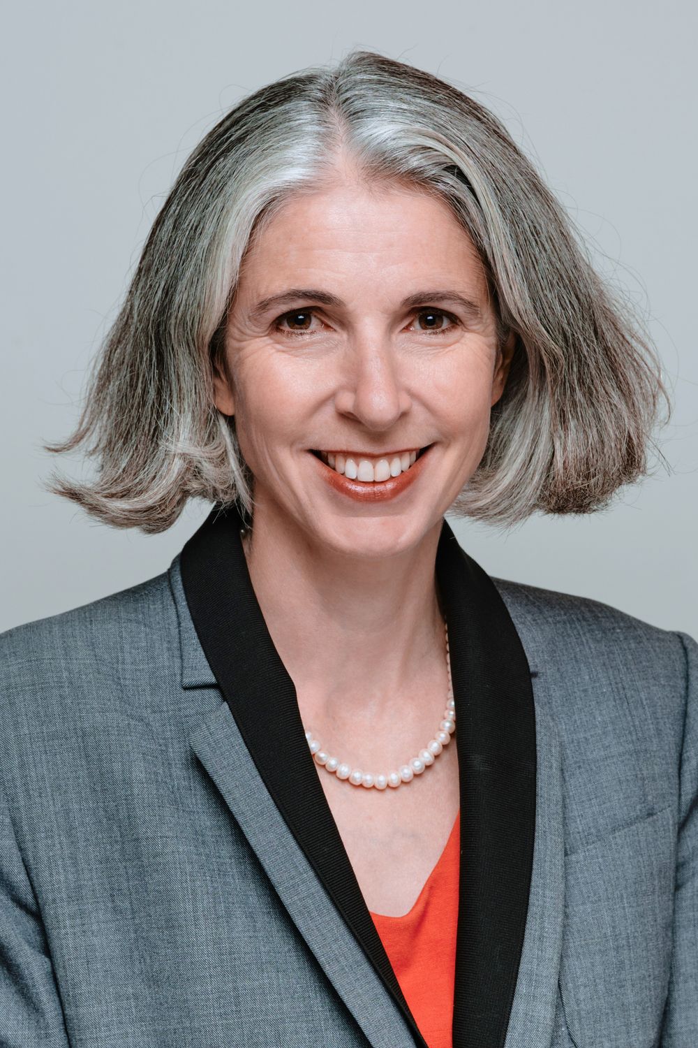 Professional headshot of a person with silver hair wearing a gray blazer, black lapels, orange top and pearl necklace.