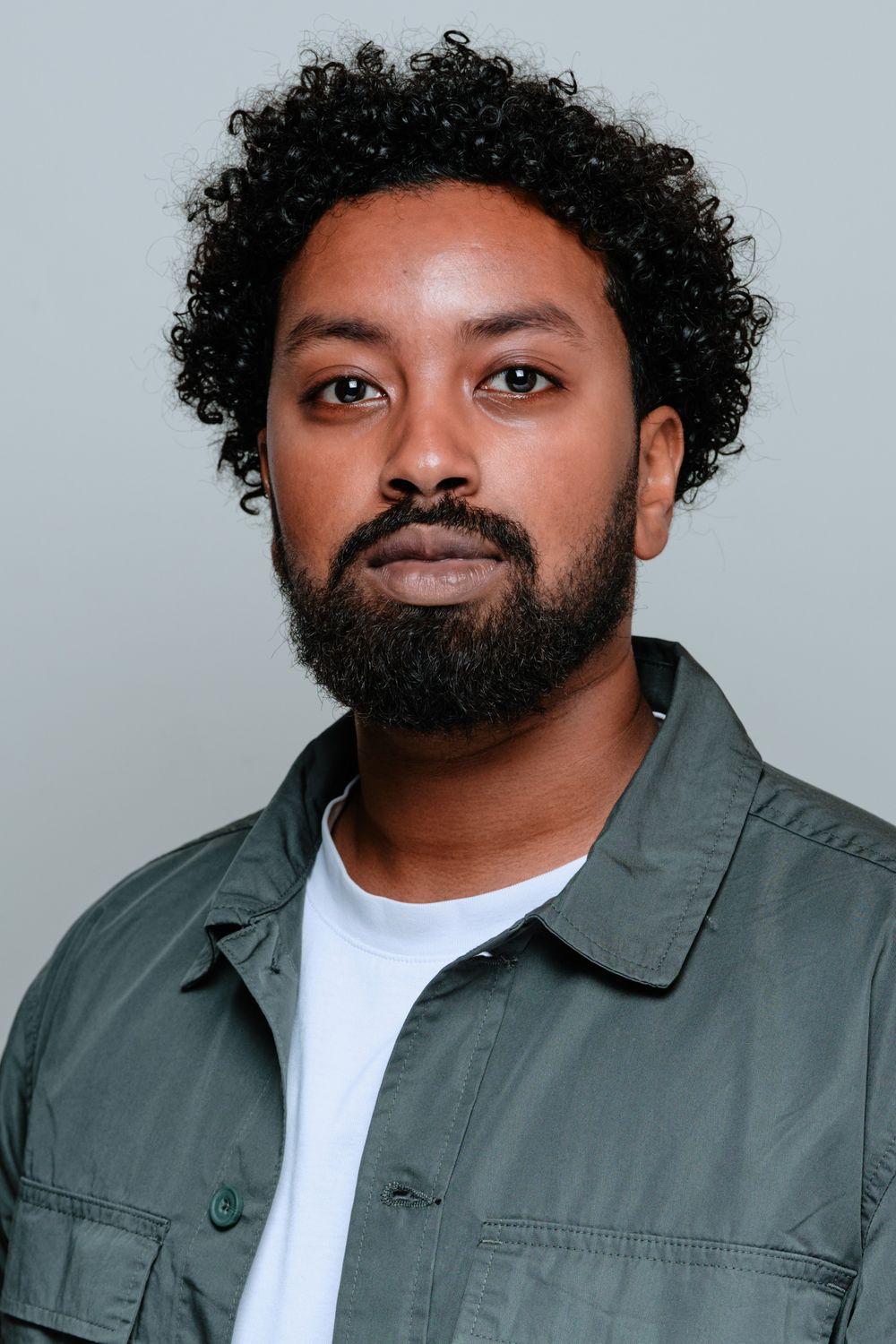 A person with curly dark hair and facial hair wearing a white t-shirt and olive green jacket poses in a photo series.
