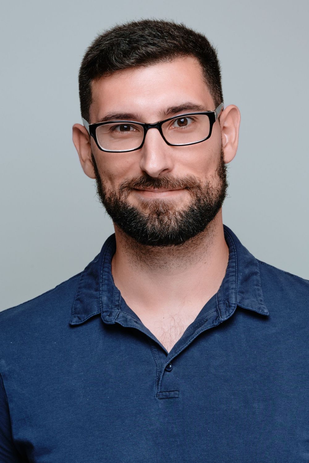 A person wearing glasses and a navy blue collared shirt poses against a light gray background.